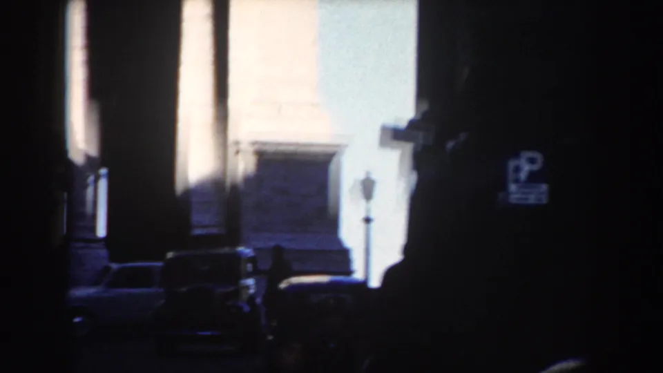 A dark Rome street opens toward sunlit stone architecture, with parked cars and small pedestrian figures visible in the shadows.