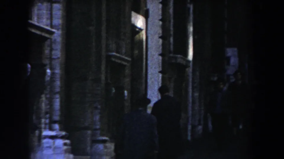 A dim Rome street scene shows tall stone facades, window frames, and pedestrians standing or walking along the shaded sidewalk.