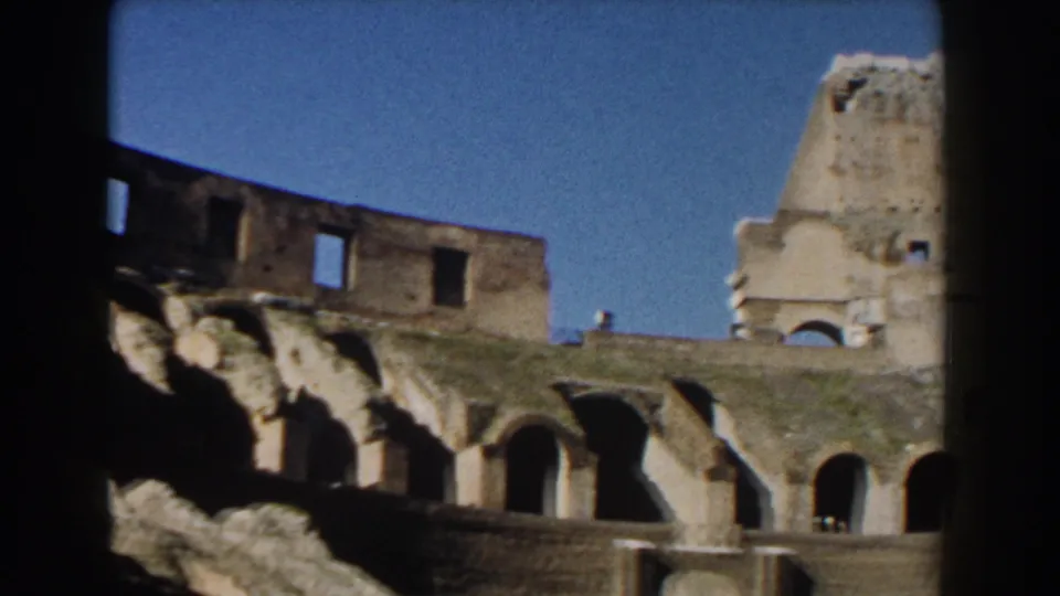 1959 8mm view of the Colosseum upper arches in Rome, with worn masonry, blue sky, and dark gate framing.