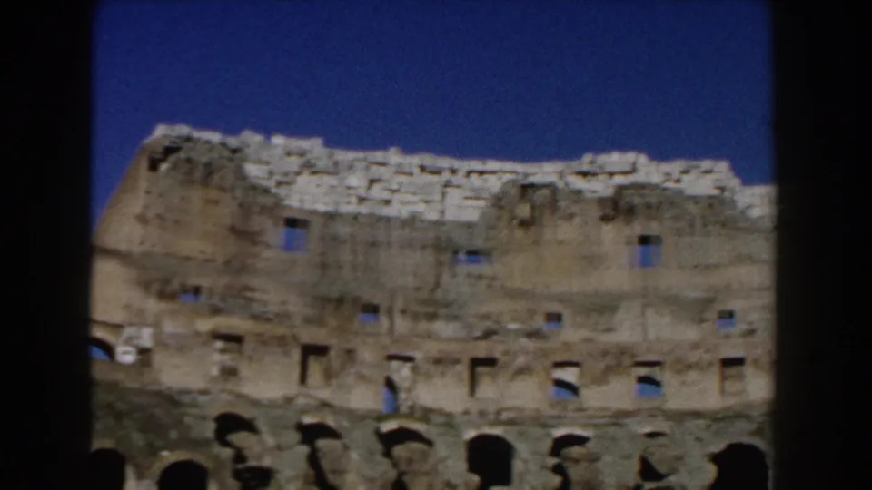 1959 8mm frame of the Colosseum in Rome, showing upper masonry, small openings, lower arches, and a distant visitor.