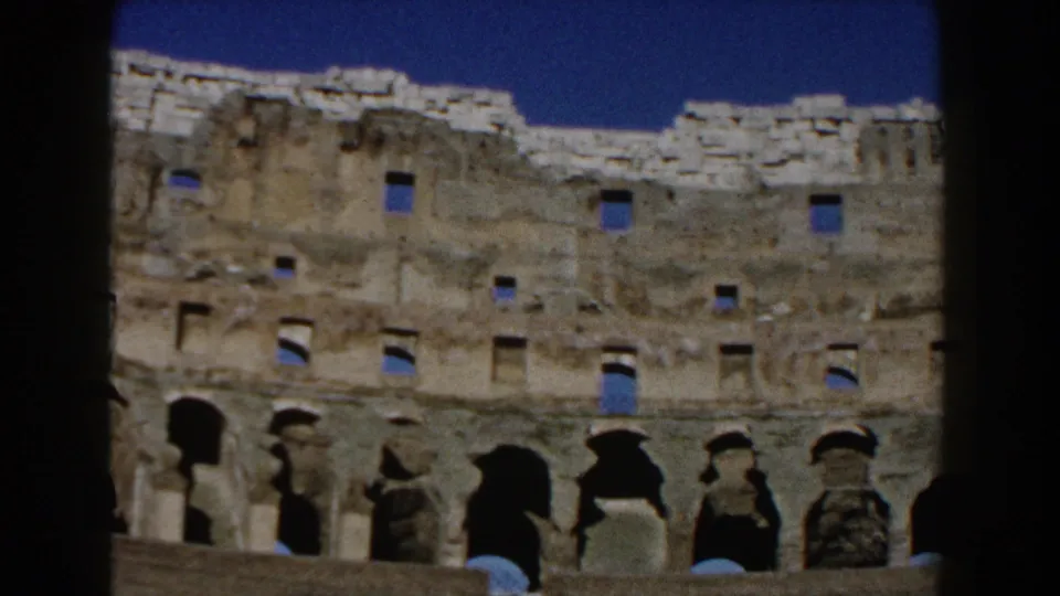 Late-1950s home-movie view of the Colosseum in Rome, with upper stonework, repeated arches, blue sky, and dark scan borders.