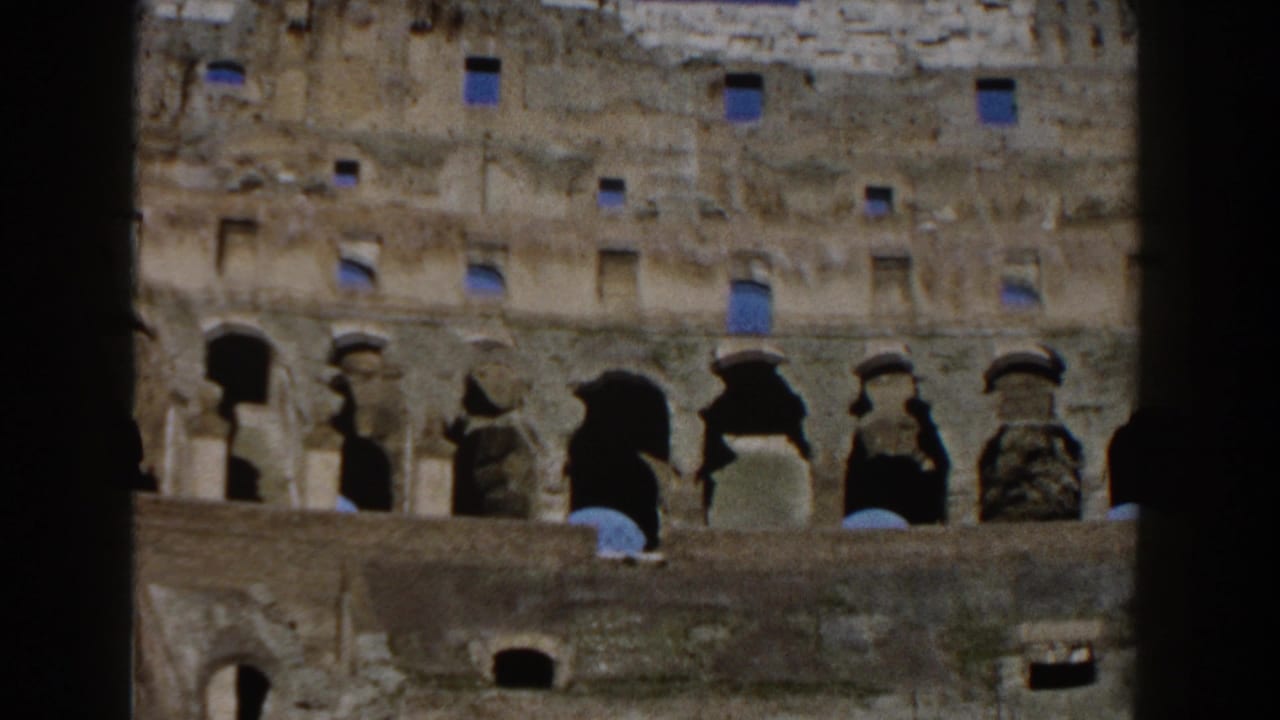 1959 8mm view of the Colosseum facade in Rome, framed by dark film edges and showing worn stone arches under blue sky.