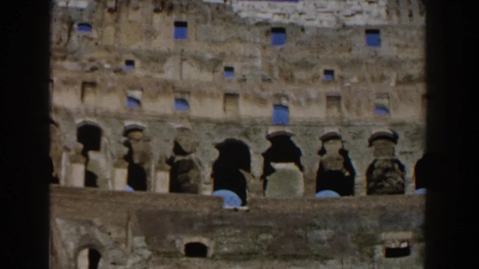 1959 8mm view of the Colosseum facade in Rome, framed by dark film edges and showing worn stone arches under blue sky.