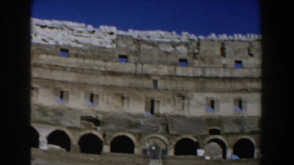 A slightly tilted 1959 8mm view of the Colosseum shows rows of arches, small upper windows, weathered stone, and clear blue sky.