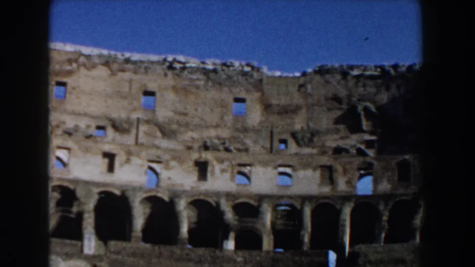 1959 home-movie frame of the Colosseum interior, with tiered arches, rough masonry, and shadowed openings beneath blue sky.