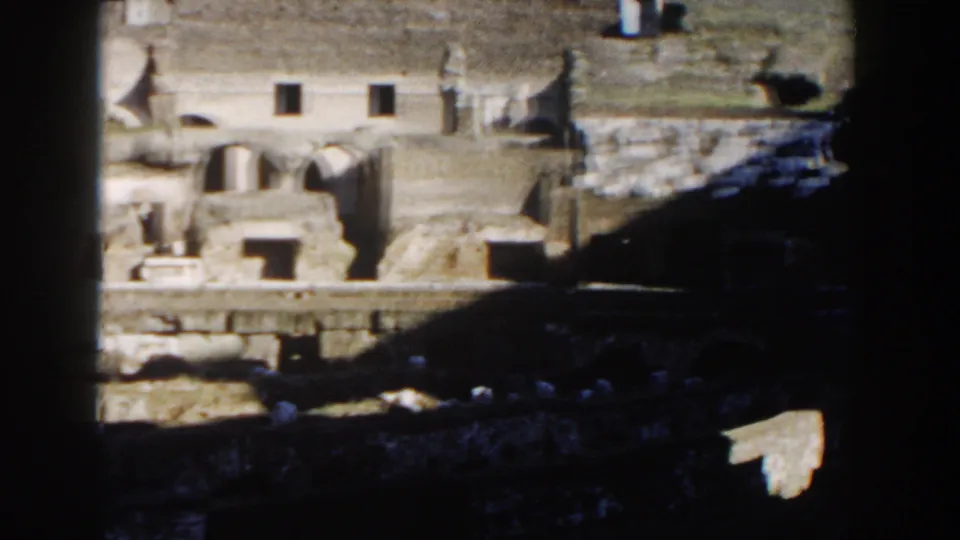 1959 home-movie frame of the Colosseum interior, showing pale stone walls, small openings, grass, and dark foreground shadow.