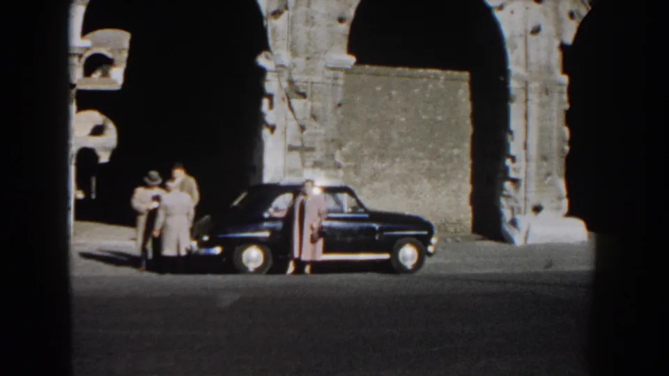 Adults in coats stand by a dark mid-century sedan near the Colosseum arches in Rome, 1959.