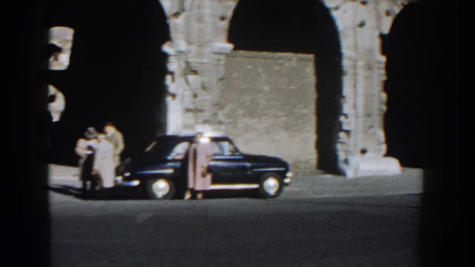Rome tourists stand close together near a dark sedan against the Colosseum's arched stone facade.