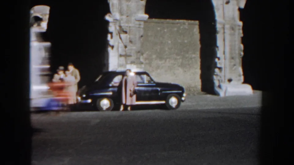 A blurred passerby or vehicle crosses the frame as adults stand beside a dark sedan near the Colosseum in Rome.
