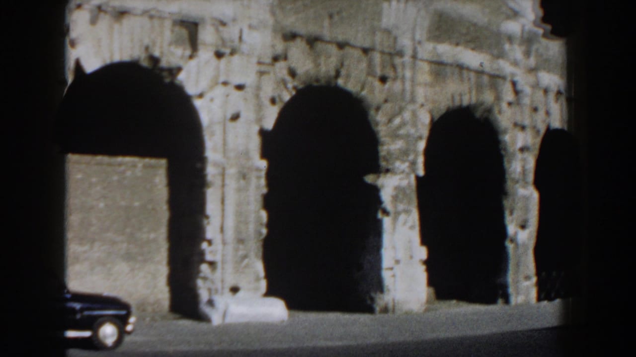 1959 Rome home-movie frame showing one side of the Colosseum with shadowed arches and a partial dark car at the edge.