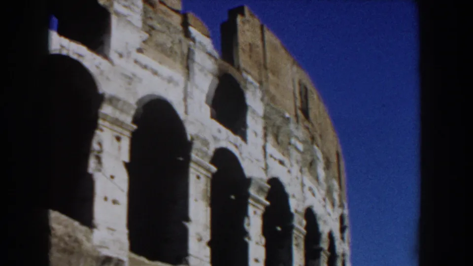 1959 Rome home-movie view of the Colosseum from below, with curved stone walls and blue sky behind it.