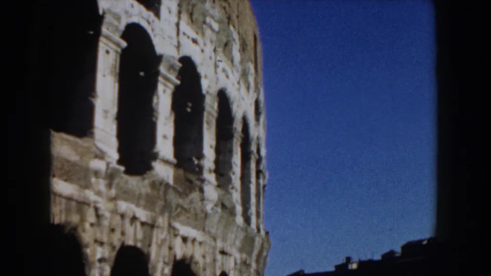 1959 Rome frame showing the Colosseum's exterior side, with bright stone, dark arches, and open blue sky.