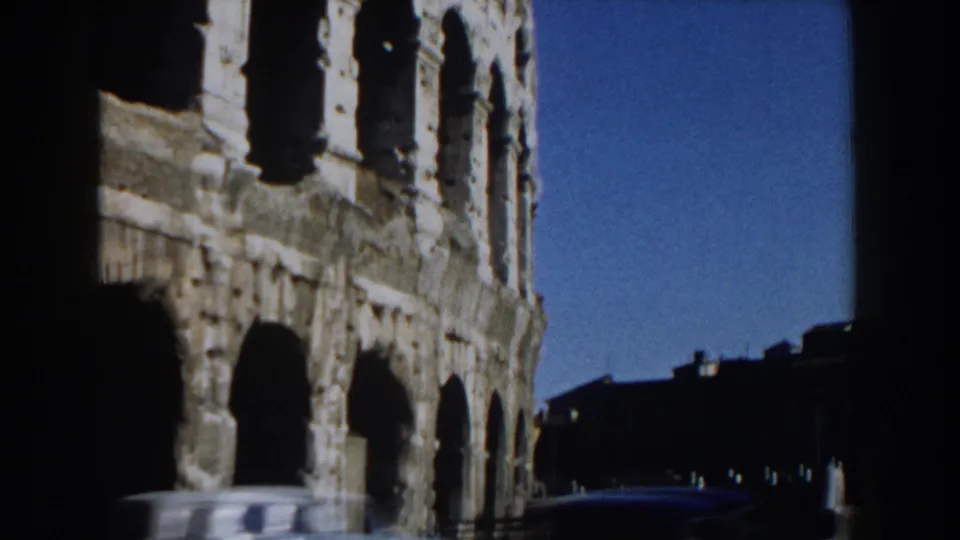 1959 home-movie frame of the Colosseum exterior in Rome, partly obscured by dark film borders and a passing car.
