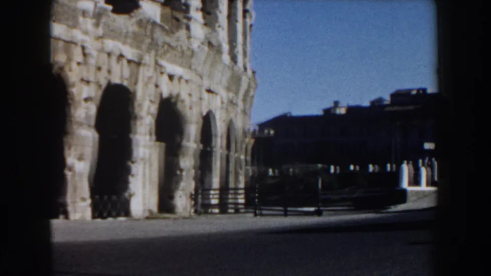 Rome Colosseum exterior appears beside a dark film-frame edge, with plaza pavement and distant buildings visible.