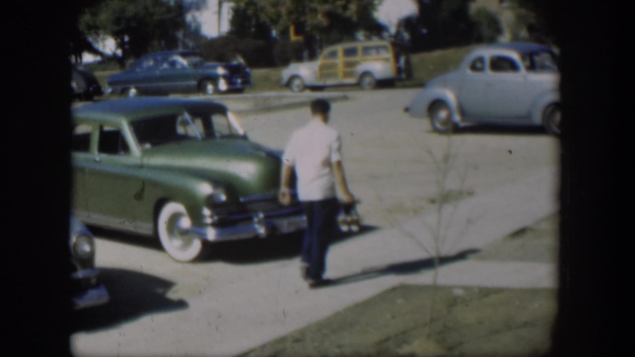 In 1954 Nevada home-movie footage, a man in a white shirt walks along a sidewalk beside a green sedan and other parked cars.