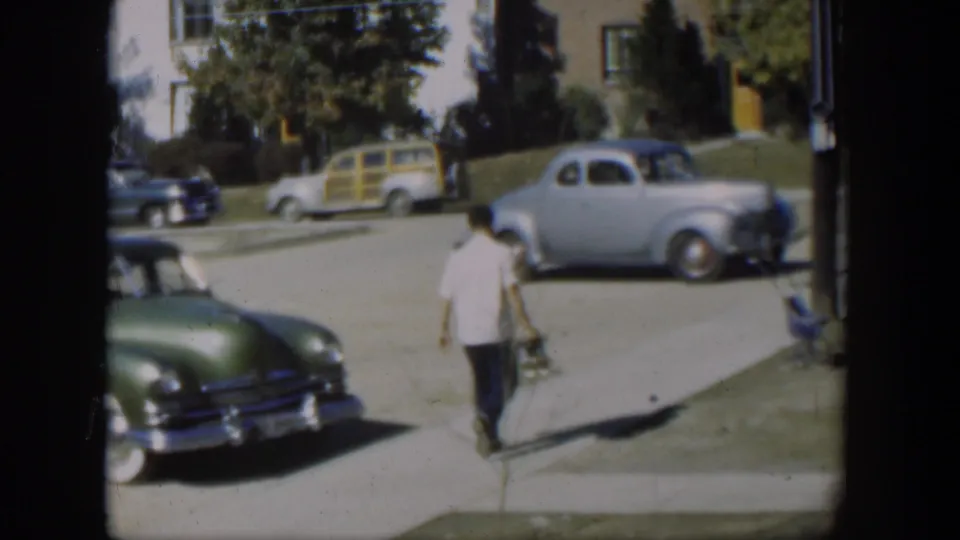 The man walks farther along the Nevada sidewalk, with a green sedan in the foreground and a light blue coupe across the street.
