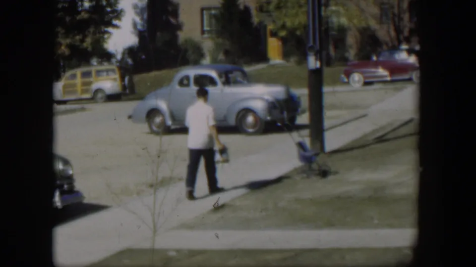 The man appears to slow or turn on the sidewalk, with a light blue coupe, wood-sided station wagon, and red car visible nearby.