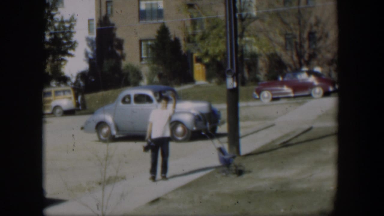 In a 1954 Nevada residential street scene, a man waves near a light blue coupe, a maroon sedan, a sidewalk, and apartment buildings.