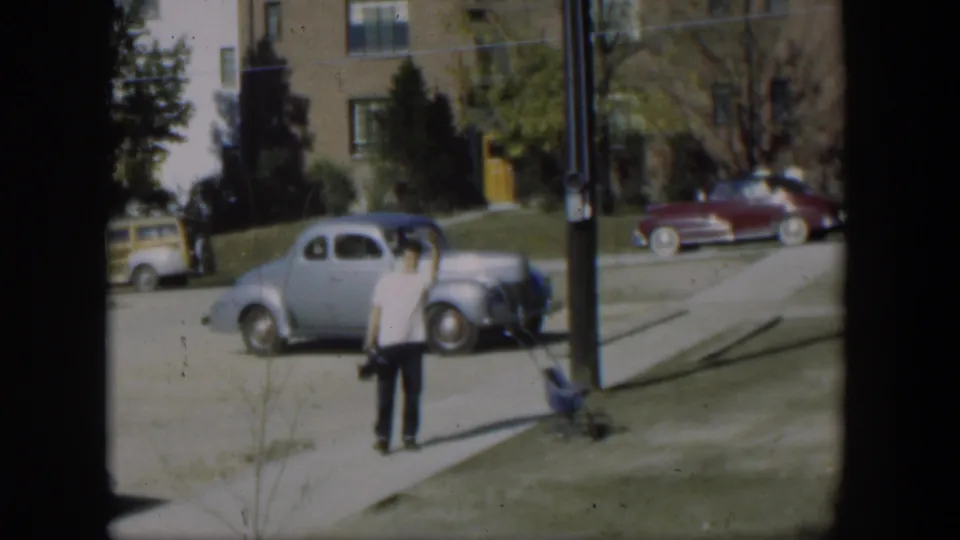 In a 1954 Nevada residential street scene, a man waves near a light blue coupe, a maroon sedan, a sidewalk, and apartment buildings.