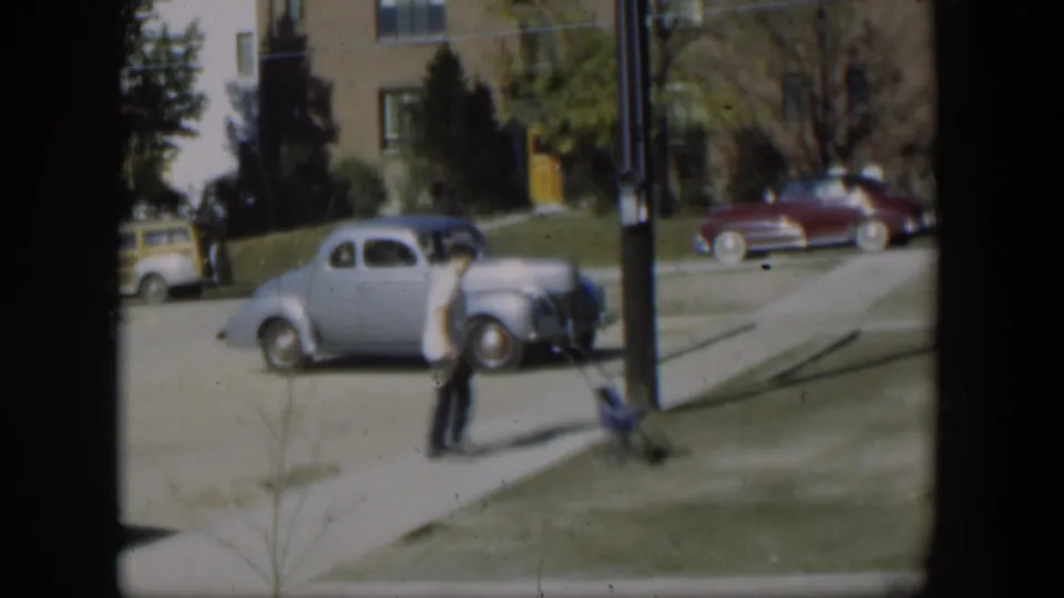 The man turns away on the sidewalk in the Nevada home-movie scene, with vintage cars, a utility pole, lawn, and apartments behind him.