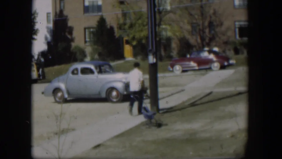 The man continues down the sidewalk in 1954 Nevada, passing a light blue coupe, maroon sedan, grass lawn, and brick apartment building.
