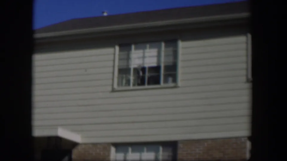 1954 Nevada 8mm frame with a closer view of a pale-sided house wall, upper window, roof edge, brick lower section, and blue sky.