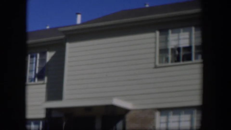 1954 Nevada home-movie frame showing the house facade with roofline, chimney, pale siding, window, brickwork, and a small entry roof.