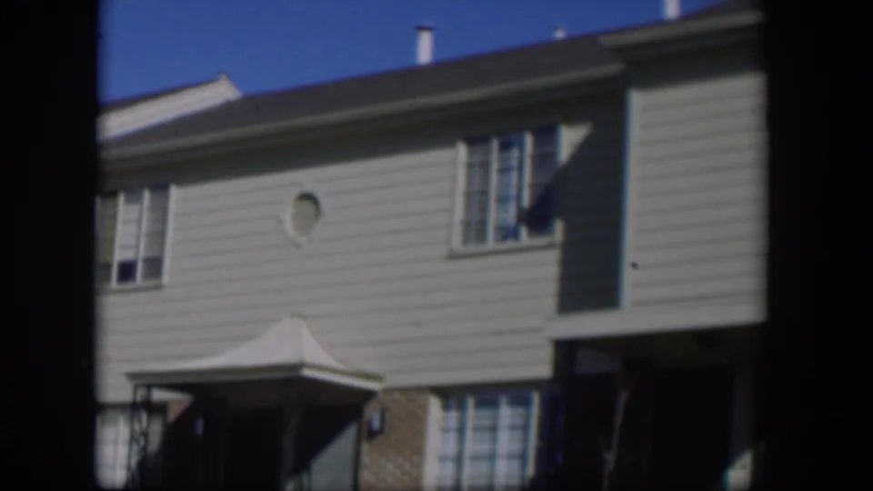 1954 Nevada 8mm frame showing the covered entry, brick lower wall, upper windows, and light horizontal siding of a suburban house.
