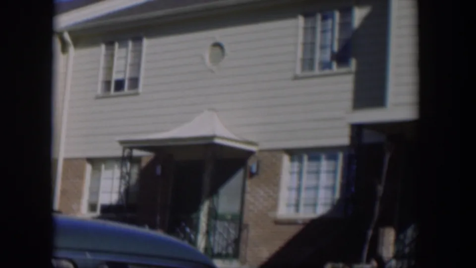 1954 Nevada home-movie frame with the house entry canopy, brick accents, windows, siding, and the top of a parked car at the bottom edge.
