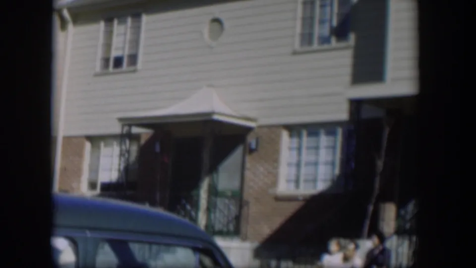 1954 Nevada home-movie frame showing a townhouse entrance, windows, brick accents, and a blue car in the foreground.