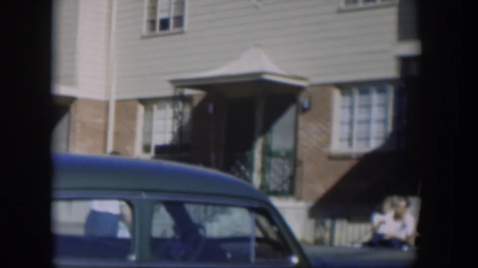 1954 Nevada home-movie view of a townhouse facade, a foreground blue car, and a person seated near the steps.