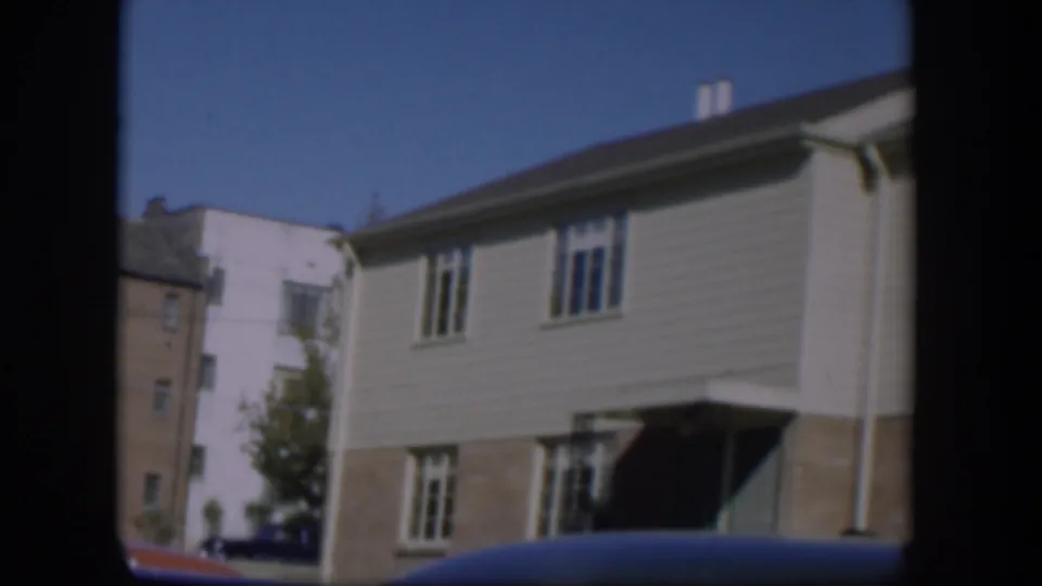 1954 Nevada residential street view with a two-story house, adjacent building, small tree, and car roof in the foreground.