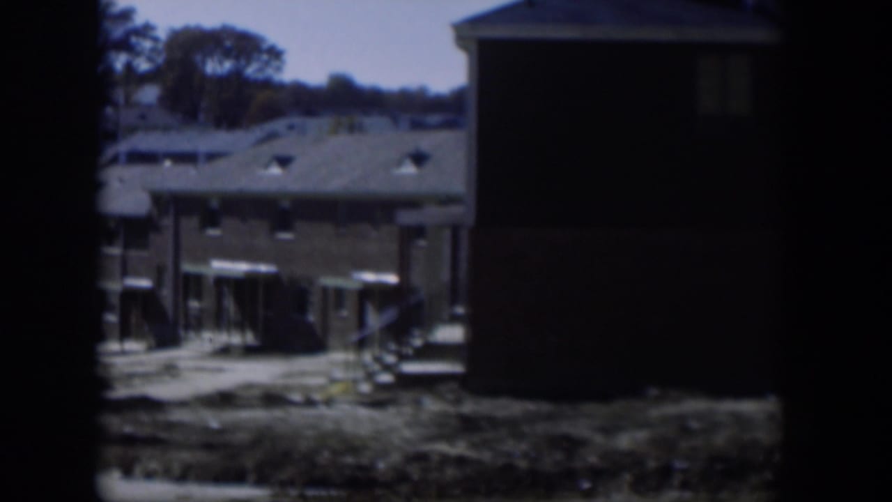 1954 Nevada 8mm frame showing low camp-style buildings, awnings, dry ground, and dark borders from the film gate.
