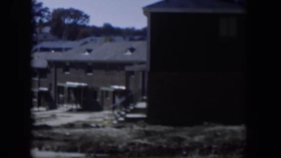 1954 Nevada 8mm frame showing low camp-style buildings, awnings, dry ground, and dark borders from the film gate.