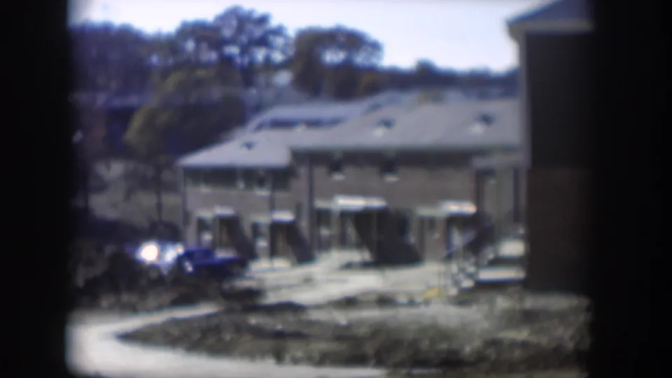 Nevada 8mm home-movie frame with low cabins, shaded awnings, a parked car, dry terrain, trees, and dark frame borders.