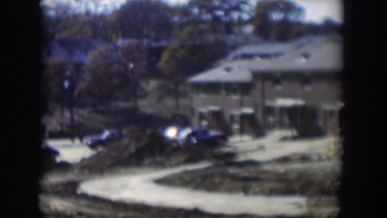 Low buildings, parked cars, dirt mounds, and a pale curved road or path.