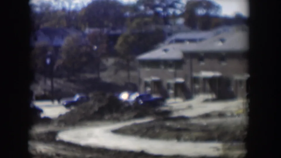 1954 Nevada home-movie frame with parked cars near low buildings, earth piles, trees, and a curved light-colored roadway or path.
