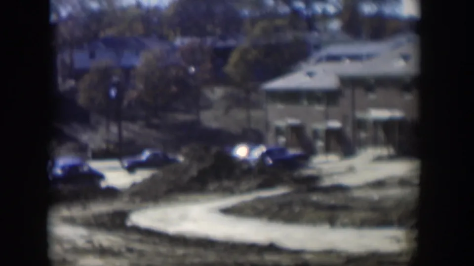 1954 Nevada 8mm frame showing classic cars along a dirt road, a low building, trees, and rough graded ground.