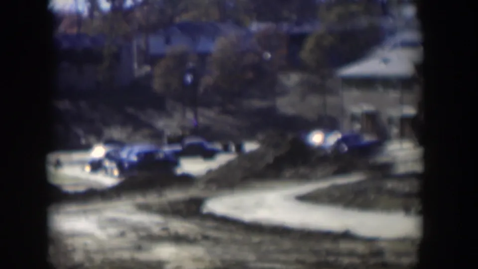 1954 Nevada home-movie frame with parked cars, a low building, trees, dirt mounds, and a curved pale road or path.