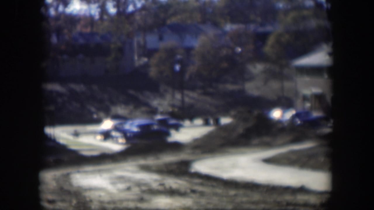 1954 Nevada home-movie frame showing cars near an apartment complex driveway, with trees, pavement, and soft 8mm exposure.