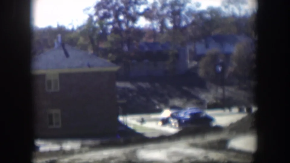 Soft 8mm frame of a 1954 Nevada apartment exterior with a blue car, pale pavement, trees, and dark film-frame edges.