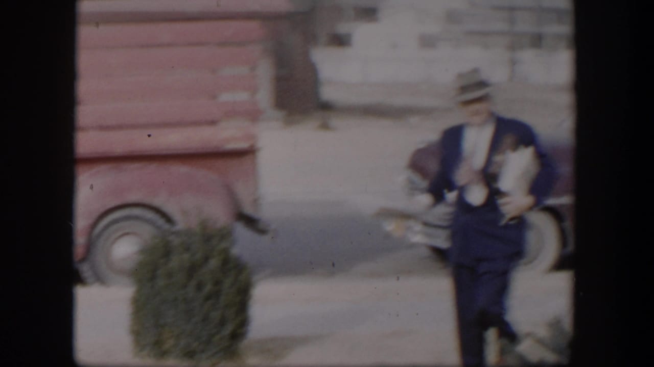 Man in blue suit and fedora walking near parked vintage vehicles.