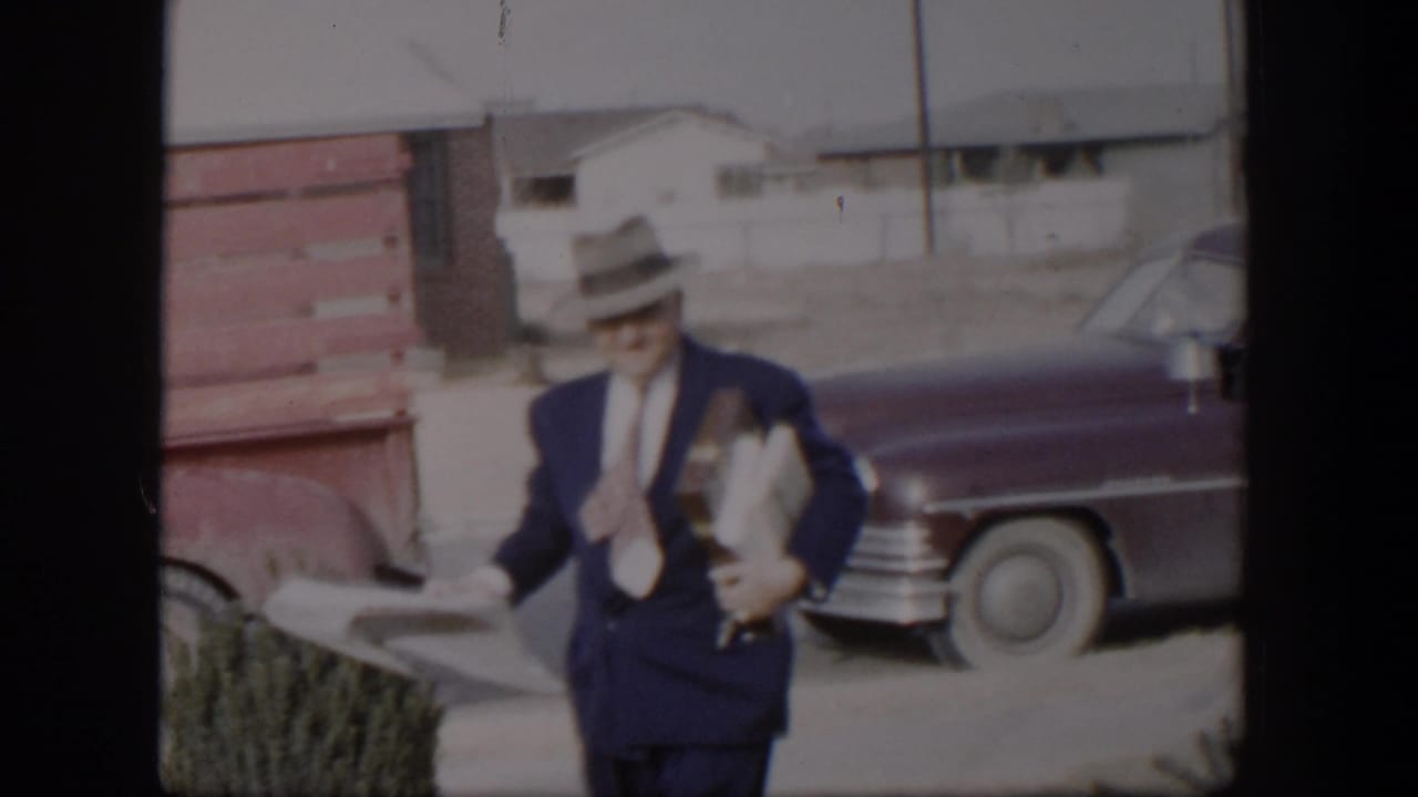 Suited man carrying papers and long packages beside vintage cars.