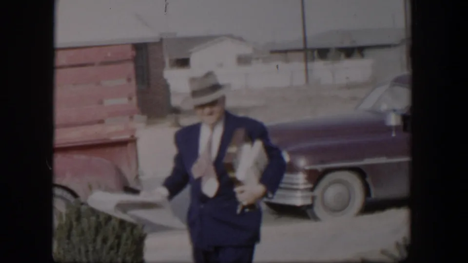 The man in a fedora carries folded papers and long packages past a pickup truck and a dark car in 1954 Nevada footage.
