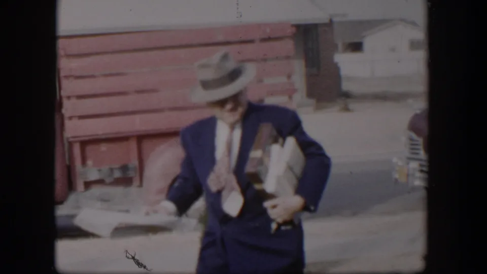 A formally dressed man in 1954 Nevada appears to look down at folded papers while carrying packages near parked cars.