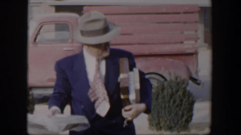 A closer 1954 Nevada frame shows the suited man in a fedora holding papers and packages beside a pink pickup and shrub.