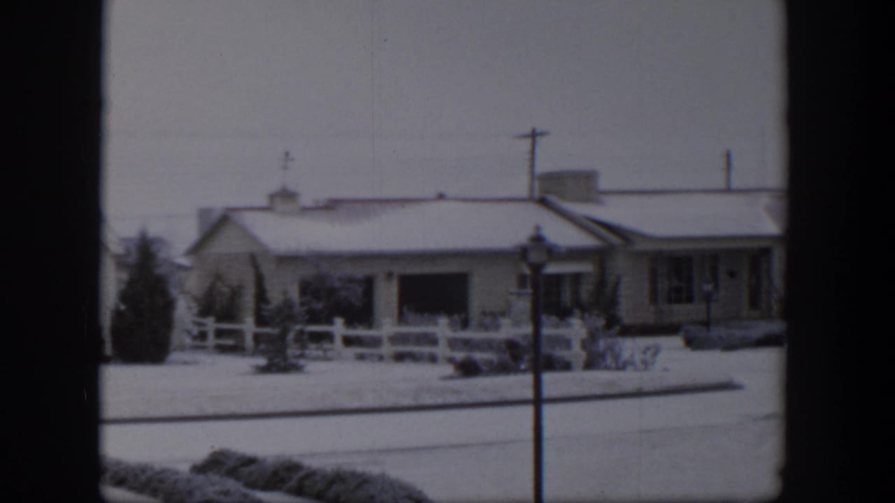 1954 Nevada home-movie frame showing a snow-covered house, white fence, streetlamp, and dark window frame edges.