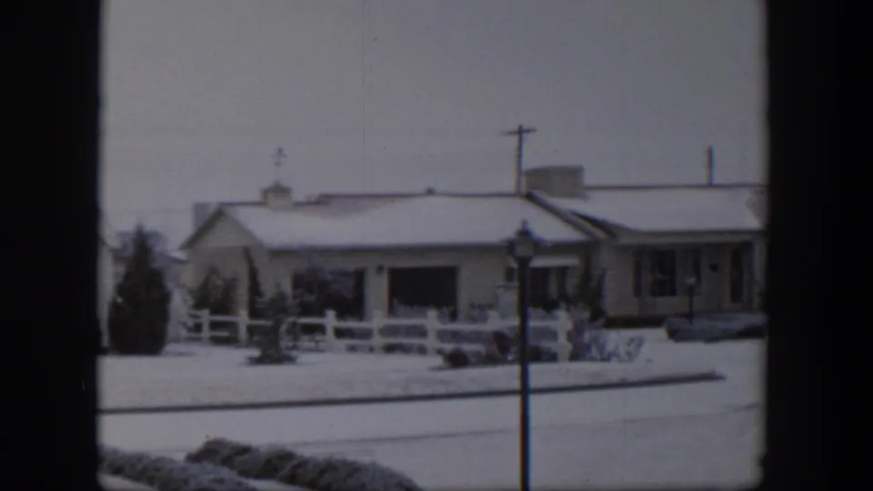 1954 Nevada home-movie frame showing a snow-covered house, white fence, streetlamp, and dark window frame edges.