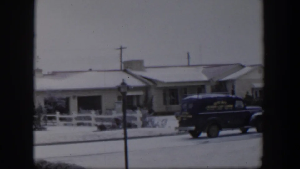 A blue delivery-style truck drives past a snow-covered house and fence in a 1954 Nevada residential scene.