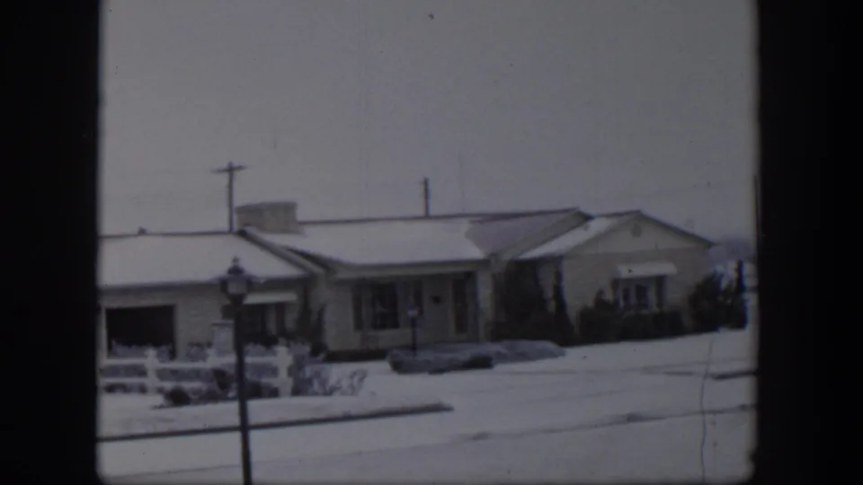 1954 Nevada 8mm frame showing ranch-style houses, white fencing, bare shrubs, and snow across the street and lawns.